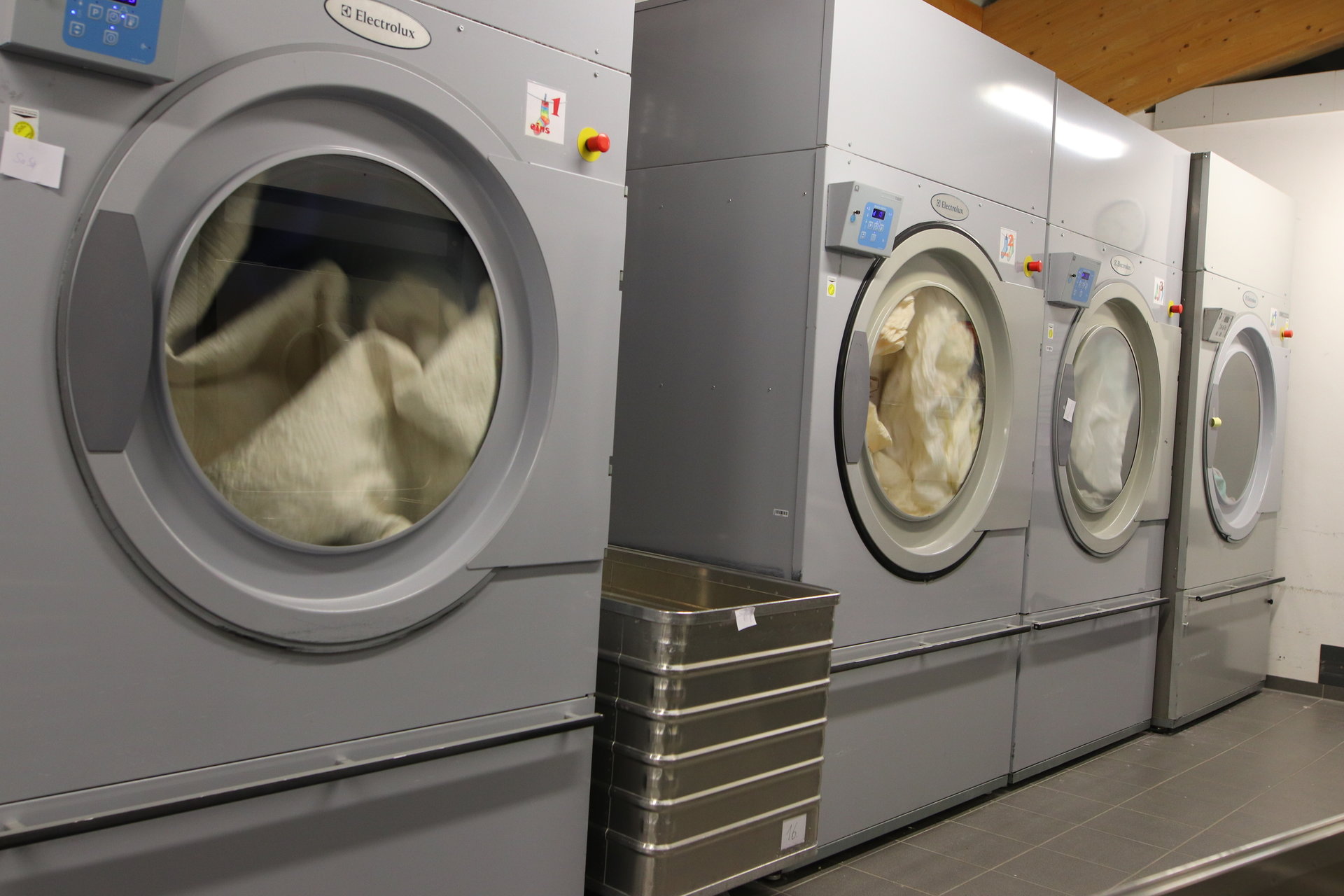 Eine Reihe grauer Electrolux-Waschmaschinen mit sichtbaren Falten im Gewebe steht in einem Waschsalon, begleitet von einem Metalleimer und einem Holzbalken über dem Kopf.